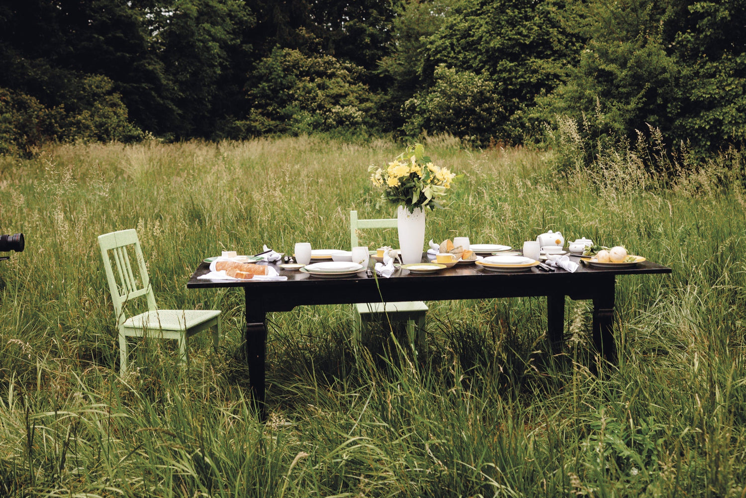 In einer hohen Sommerwiese steht ein gedeckter Tisch mit Porzellanservice der Porzellan Manufaktur Nymphenburg. Das Arrangement mit Kuchen, Croissants, Käseplatte und einer Blumenvase wirkt wie eine nostalgische Einladung zu einem Picknick im Grünen – poetisch, reduziert und zugleich detailverliebt inszeniert.