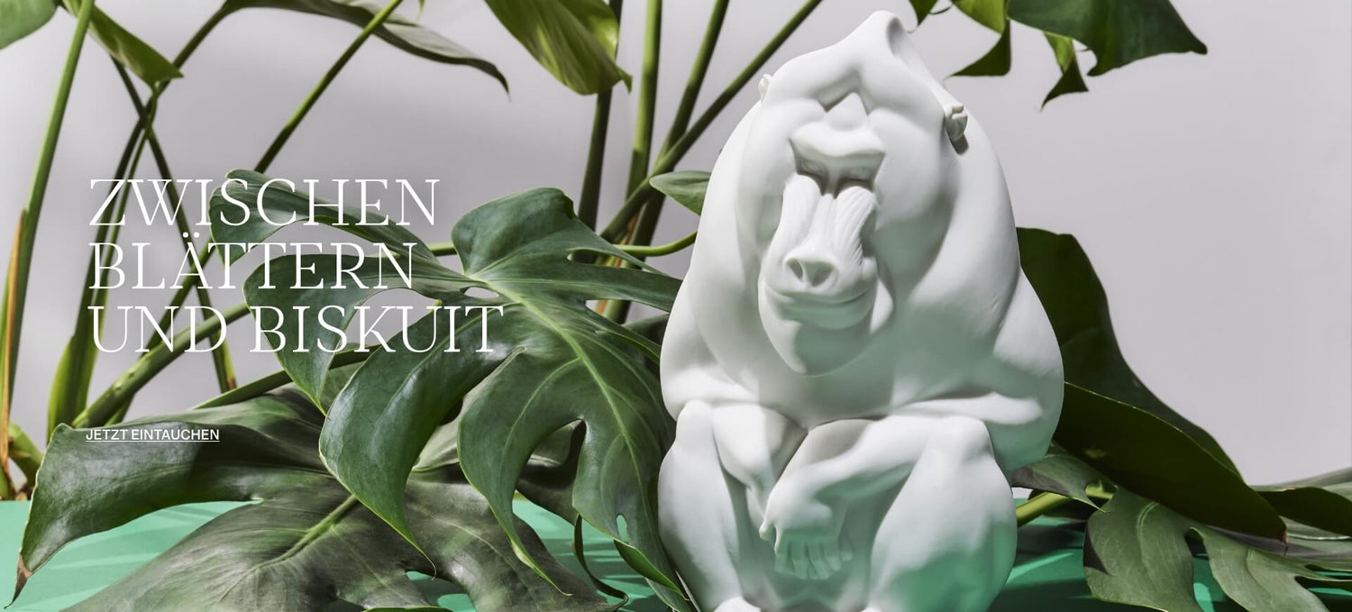 Weiße Biskuitporzellanfigur eines sitzenden Gorillas, inszeniert vor großflächigen grünen Monstera-Blättern. Die plastisch modellierte Skulptur steht auf einem leuchtend grünen Untergrund, flankiert von der Textzeile „ZWISCHEN BLÄTTERN UND BISKUIT“.