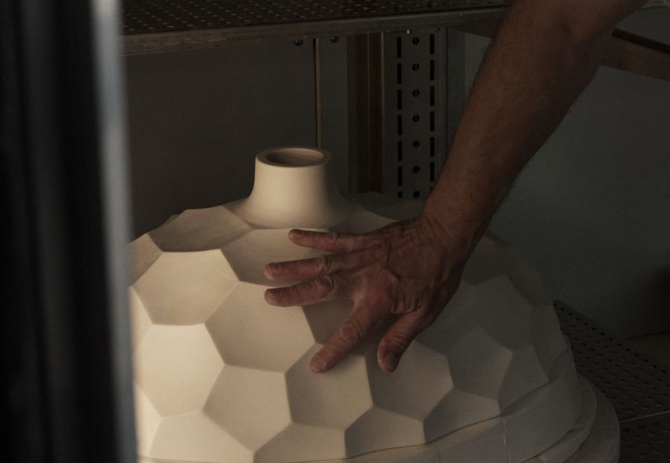 Hand eines Handwerkers stützt einen großen, unglasierten Porzellanschirm mit facettierter Wabenstruktur in einem Regal der Werkstatt.
A craftsman’s hand supports a large, unglazed porcelain shade with a faceted honeycomb structure on a workshop shelf.