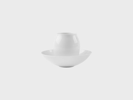 Zwei weiße Porzellanobjekte auf hellem Hintergrund: eine kleiner, bauchiger Mokka Becher steht mittig in einer flachen Schale. Beide Objekte sind schlicht, glänzend glasiert und ohne Dekor.
Two white porcelain objects on a light background: a small, rounded mocca beaker stands centered inside a shallow bowl. Both pieces are simple, glossy glazed, and undecorated.