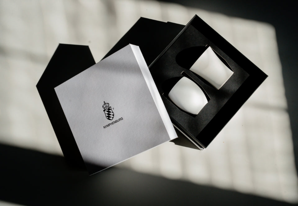 Geöffnete Nymphenburg Geschenkbox mit zwei weißen Porzellanbechern in schwarzer Einlage.
Open Nymphenburg gift box with two white porcelain cups in a black insert.