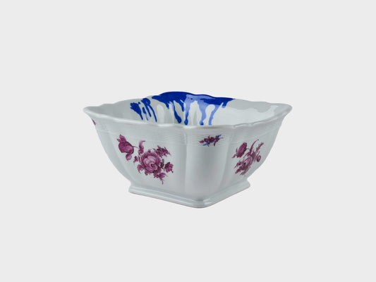 Viereckige weiße Porzellanschale mit geschwungenem Rand, dekoriert mit violetten Blumenmotiven und blauen Farbtropfen im Inneren.
Square white porcelain bowl with a scalloped rim, decorated with purple floral motifs and blue paint drips inside.