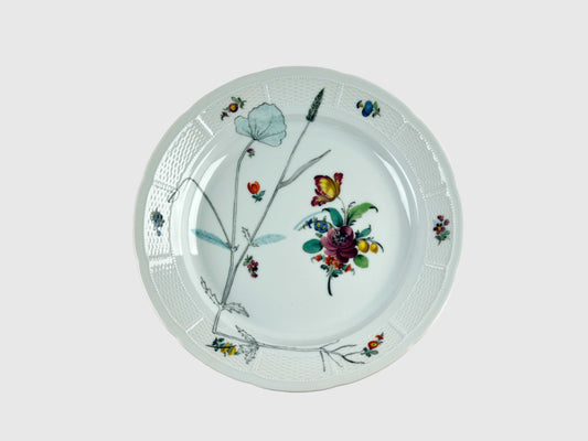 Runde Platte mit leicht gewelltem Rand und feiner Korbrelief-Struktur. Die Mitte ziert ein farbenfrohes Blumenbouquet, ergänzt durch feine botanische Zeichnungen in Schwarz. Der Tellerrand ist mit kleinen, farbigen Blüten und Insekten dekoriert.
Round porcelain platter with a gently scalloped edge and fine basket-weave relief. The center features a colorful floral bouquet, complemented by delicate black botanical line drawings. The rim is adorned with small, colorful flowers and insects.
