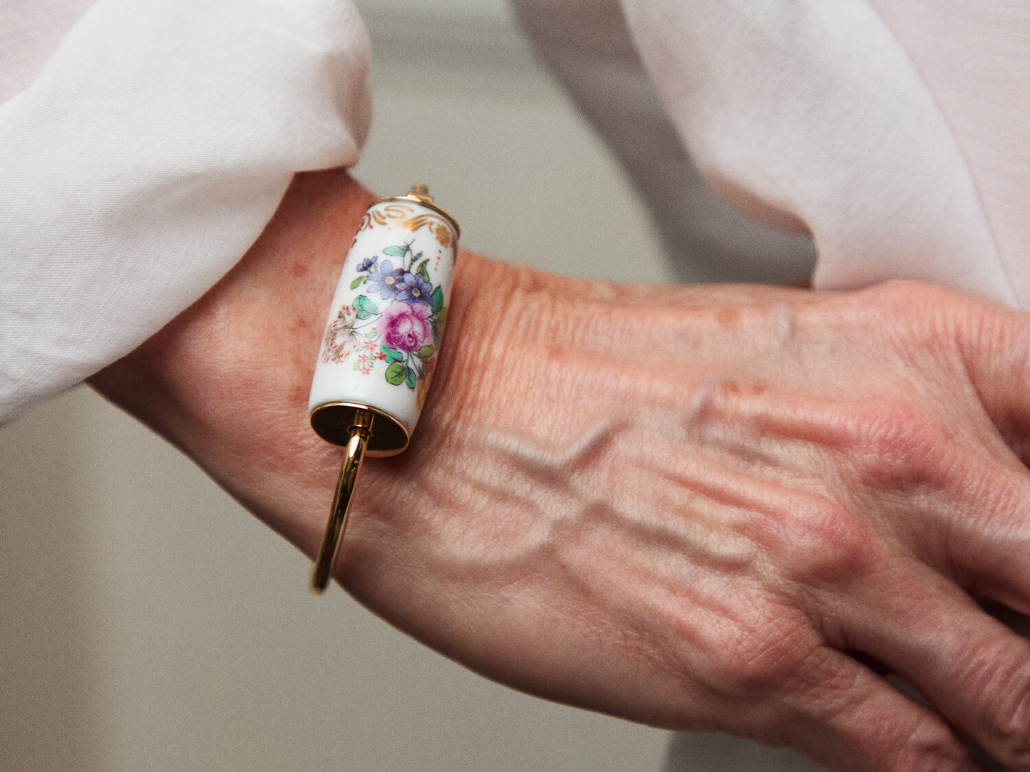 Tragefoto: Armreif mit handbemaltem Porzellan-Zylinder am Handgelenk, weiße Bluse im Hintergrund.
Lifestyle shot: bangle with hand-painted porcelain cylinder worn on the wrist, white blouse in the background.