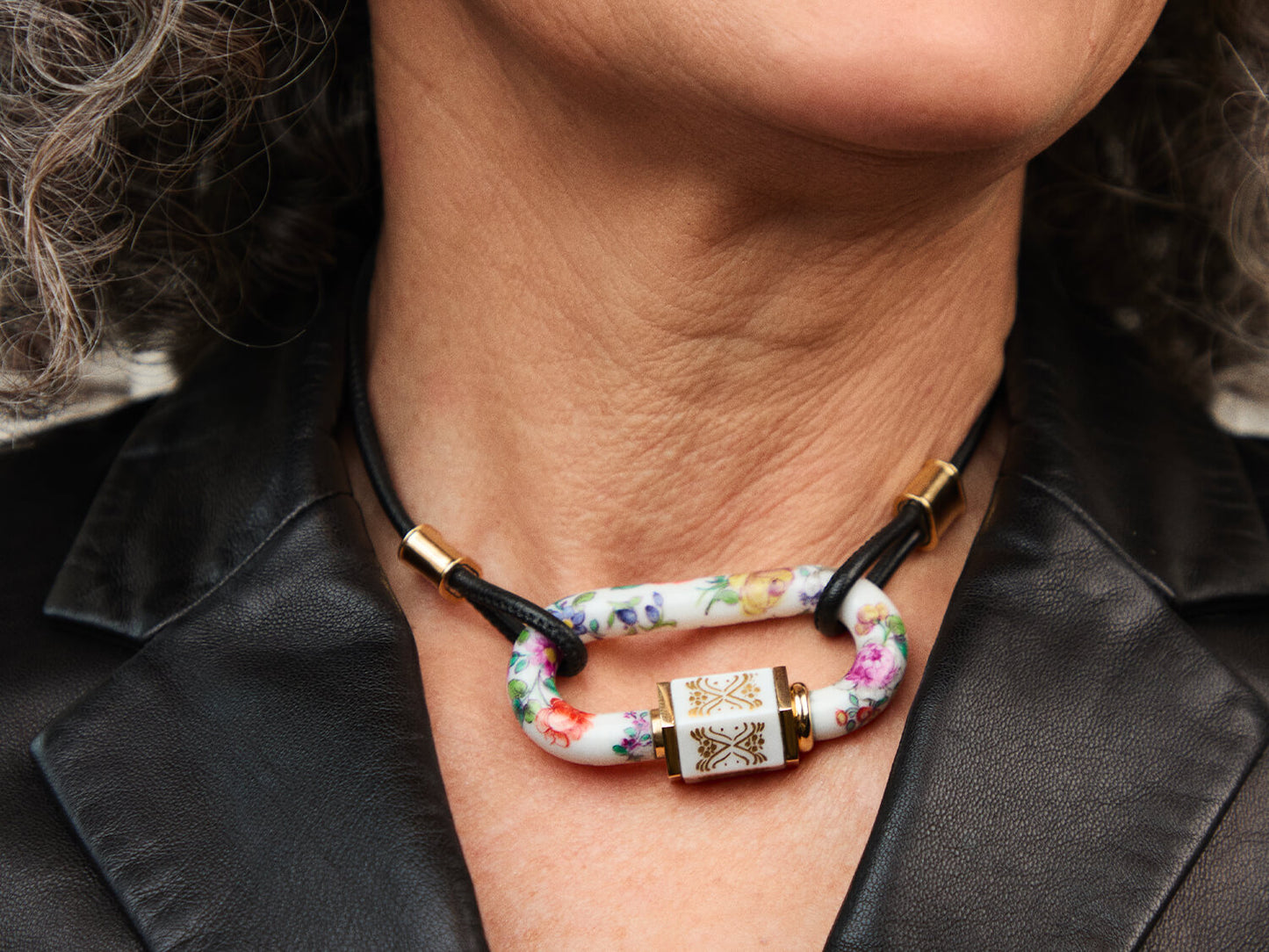 Getragenes Porzellan-Schloss am Hals – Close-up; schwarze Lederkordel, Golddetails, handbemalte Blumen auf weißem Porzellan.
Worn porcelain lock at the neck – close-up; black leather cord, gold details, hand-painted flowers on white porcelain.
