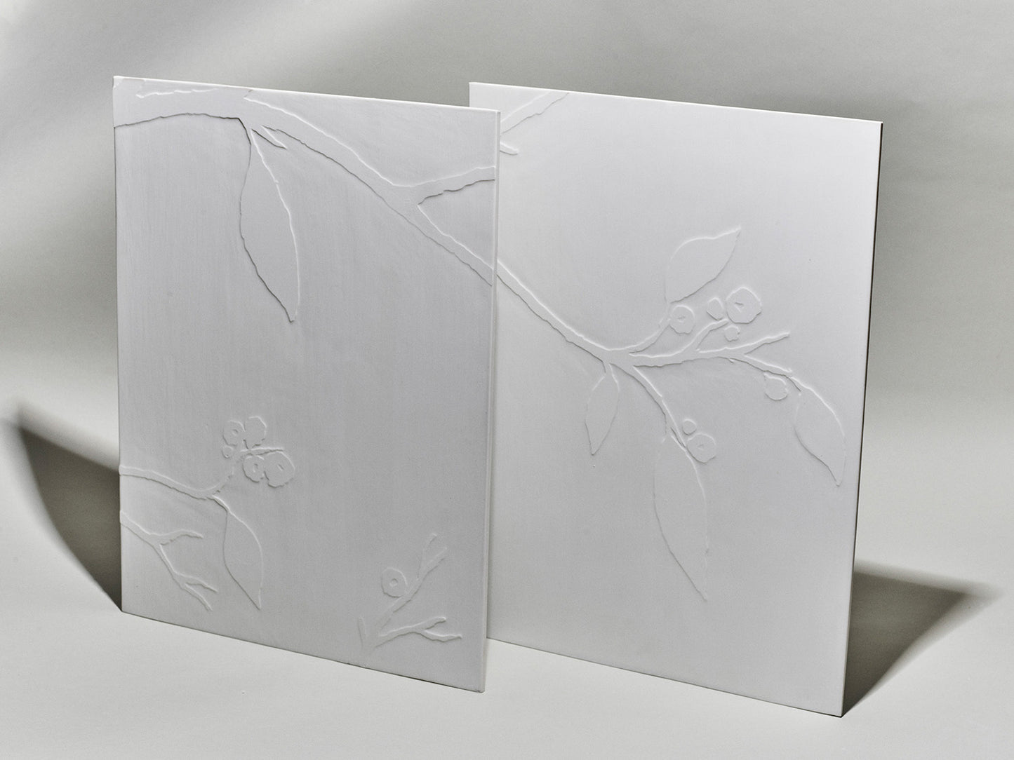 Zwei weiße Porzellanfliesen mit reliefiertem Kirschblütenzweig auf hellem Hintergrund.
Two white porcelain tiles with a relief cherry blossom branch on a light background.