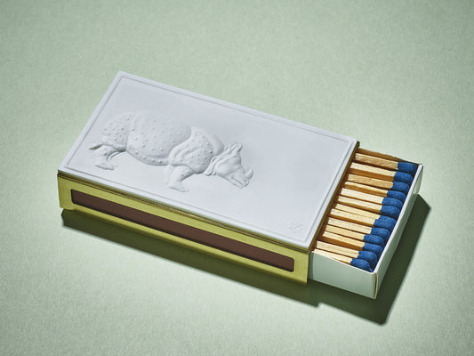 Rechteckiges Streichholzetui mit weißem Porzellanrelief eines Nashorns auf messingfarbener Hülle, blaue Zündholzköpfe sichtbar.
Rectangular matchbox case with white porcelain rhino relief on a brass-coloured sleeve, blue-tipped matches visible.