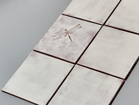 Quadratische, weiße Majolika-Fliese mit leichtem Verlauf und erhabenem Relief einer filigranen Libelle im Zentrum.
Square white-coloured majolica tile with subtle shading and a raised relief of a delicate damselfly in the centre.