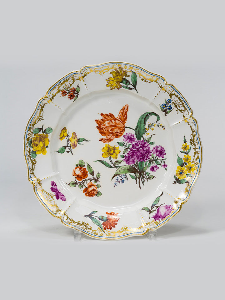 Barocker Porzellanteller mit goldverziertem Rand und bunter Blumenmalerei auf weißem Grund.
Baroque porcelain plate with a gilded rim and colourful floral painting on white background.