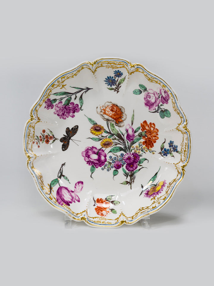 Barocker Teller mit floralem Dekor, goldverziertem Rand und schwarzem Schmetterling.
Baroque plate with floral décor, gilded rim, and black butterfly.
