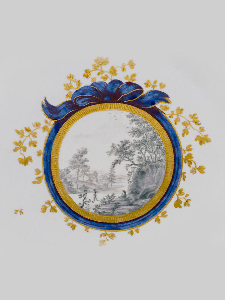 Detailansicht eines Porzellanmedaillons mit feiner Landschaftsmalerei in Schwarz-Weiß, umrahmt von kobaltblauer Schleife und Golddekor.
Close-up of a porcelain medallion with fine black-and-white landscape painting, framed by cobalt-blue ribbon and gold decoration.