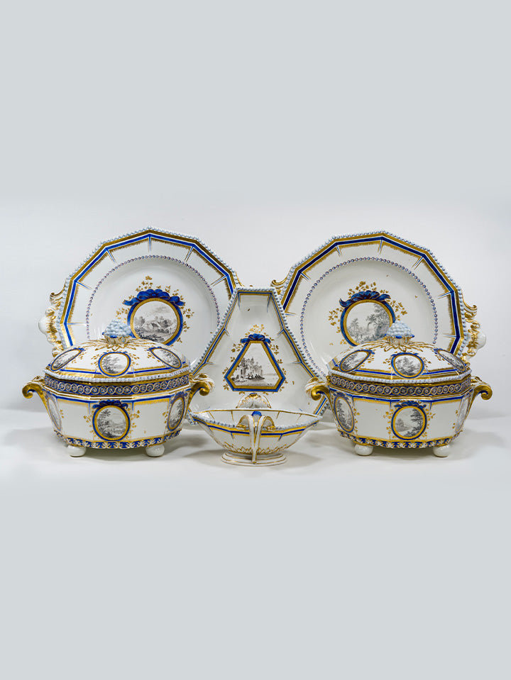 Porzellan-Ensemble aus der Serie „Perl“ mit zwei Terrinen, Platztellern, Sechskantplatte und Sauciere, verziert mit goldblauen Landschaftsmedaillons.
Porcelain ensemble from the “Perl” series featuring two tureens, charger plates, hexagonal platter and sauceboat, adorned with gold-blue landscape medallions.