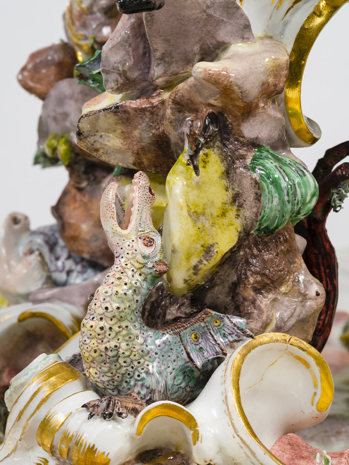 Phantasievolles, echsenartiges Tier mit aufgerissenem Maul und detailreich bemaltem Körper, modelliert zwischen Felsen und Porzellanschnecke.
Fantastic lizard-like creature with gaping mouth and intricately painted body, positioned among rocks and a porcelain snail.