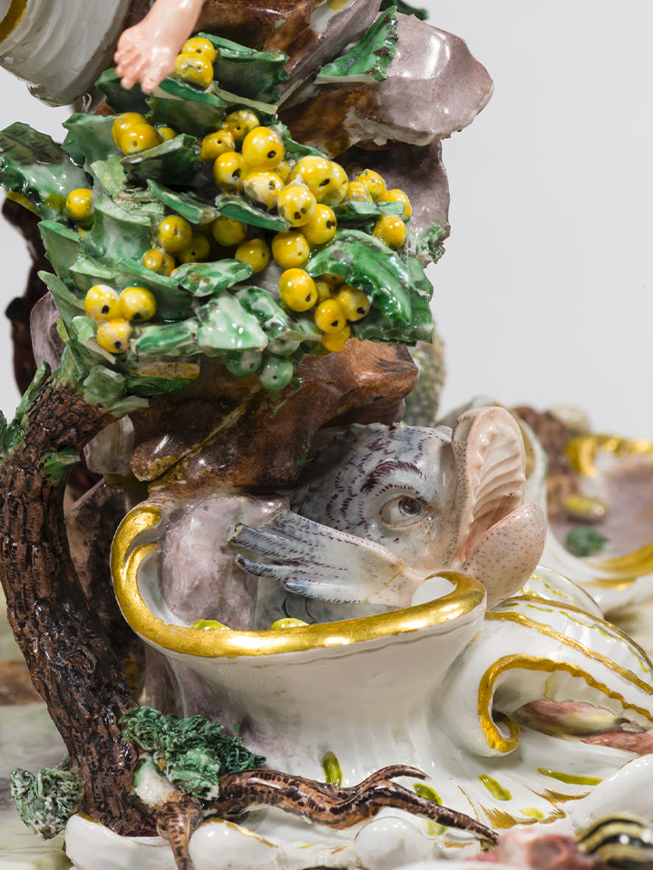 Gelbe Porzellanbeeren auf grünem Blattwerk neben einem fein modellierten Fischkopf, teils hinter Blattwerk versteckt.
Yellow porcelain berries on leafy greenery beside a delicately modelled fish head, partially hidden by foliage.