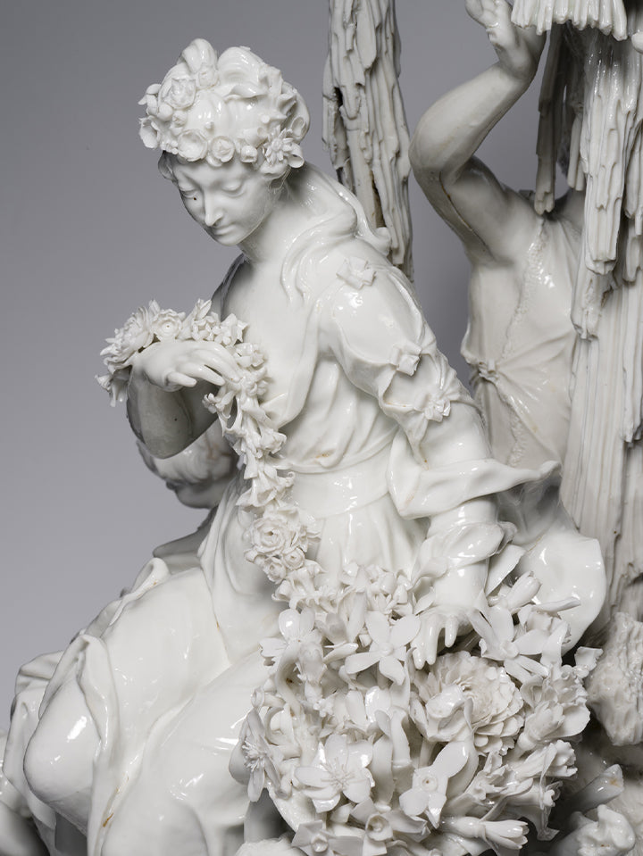 Detail einer Porzellanfigur: Eine sitzende Frau mit Blumenkranz im Haar und floraler Girlande, kunstvoll gefertigt aus weißem Biskuitporzellan.