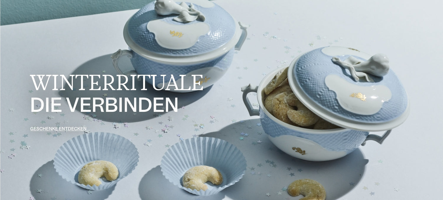 Hellblaue Porzellanschalen mit Deckel und Tierfigur, gefüllt mit Vanillekipferln, auf sternbestreutem Untergrund mit Schriftzug „Winterrituale die verbinden“.