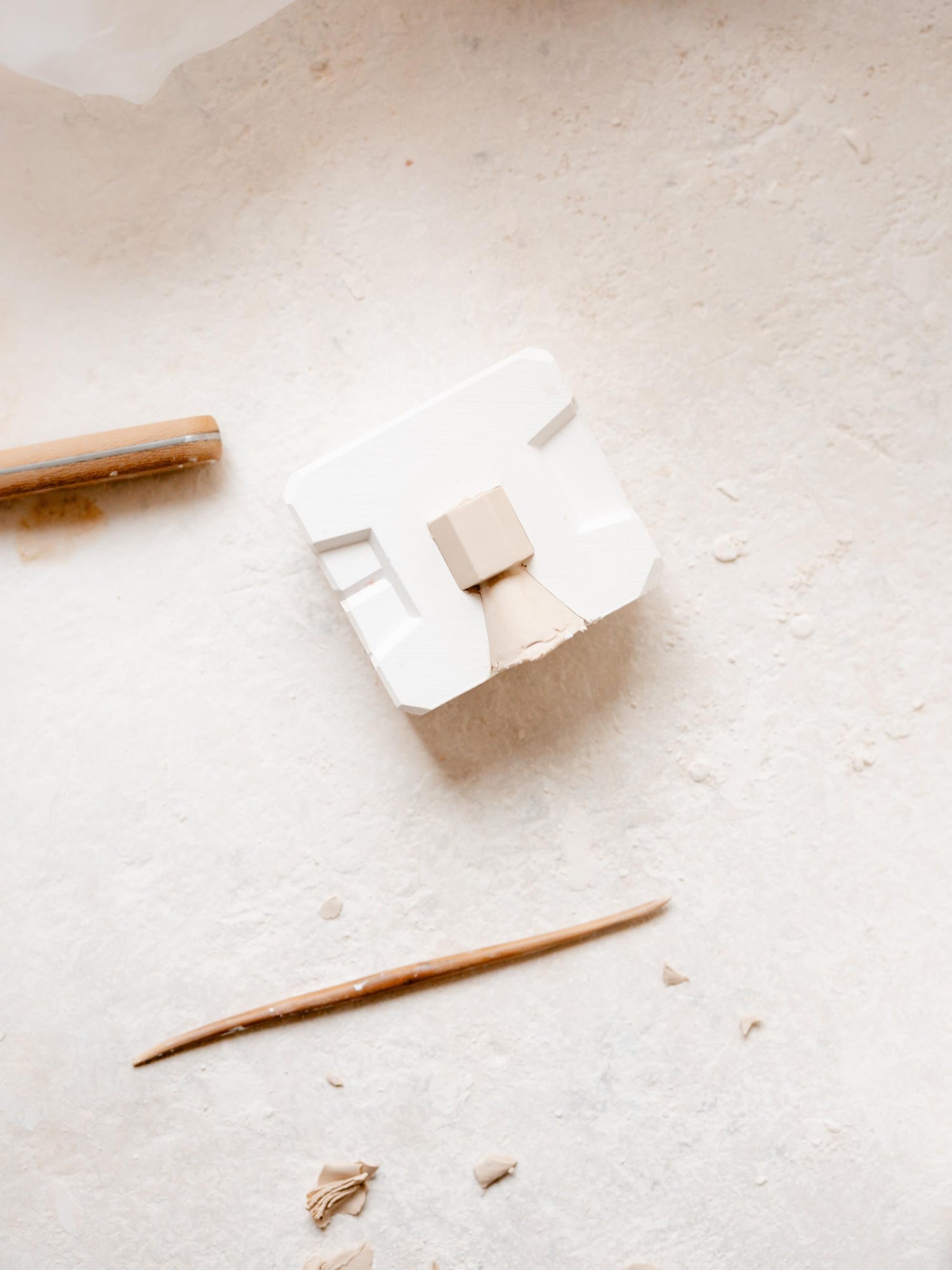 Werkstattaufnahme: kleine Gipsform mit eingefüllter Porzellanmasse auf heller Arbeitsfläche; daneben Holz-Modellierstab und Tonspäne.

Workshop scene: small plaster mould filled with porcelain slip on a light work surface; a wooden modelling tool and clay scraps nearby.