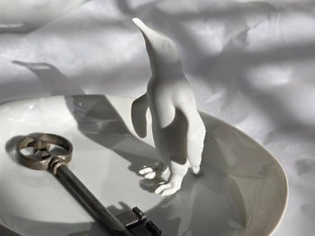 Weiße Porzellanfigur eines stehenden Pinguins auf einer ovalen Platte neben einem alten Metallschlüssel.

White porcelain figure of a standing penguin on an oval plate beside an antique metal key.