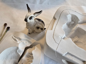 Glasierter Porzellanhase neben geöffneten Gipsformen und Pinseln auf weißem Untergrund.

Glazed porcelain rabbit beside opened plaster molds and brushes on a white surface.