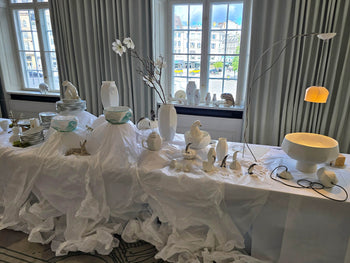 Weite Ausstellungsszene mit weißem Porzellan, leuchtender Tischlampe und dekorierten Tischen vor großen Fenstern.

Wide exhibition scene with white porcelain, a glowing table lamp, and styled tables in front of large windows.