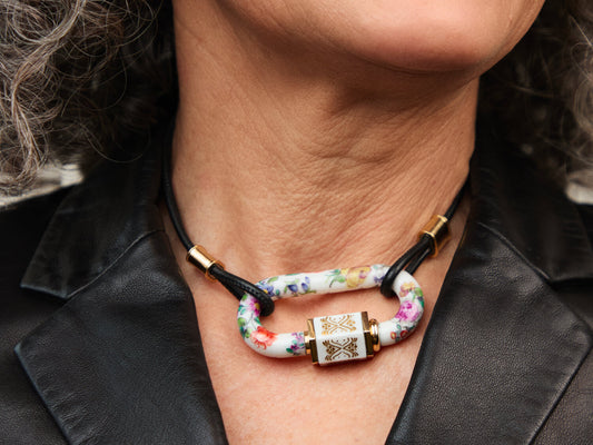 Getragenes Porzellan-Schloss am Hals – Close-up; schwarze Lederkordel, Golddetails, handbemalte Blumen auf weißem Porzellan.

Worn porcelain lock at the neck – close-up; black leather cord, gold details, hand-painted flowers on white porcelain.