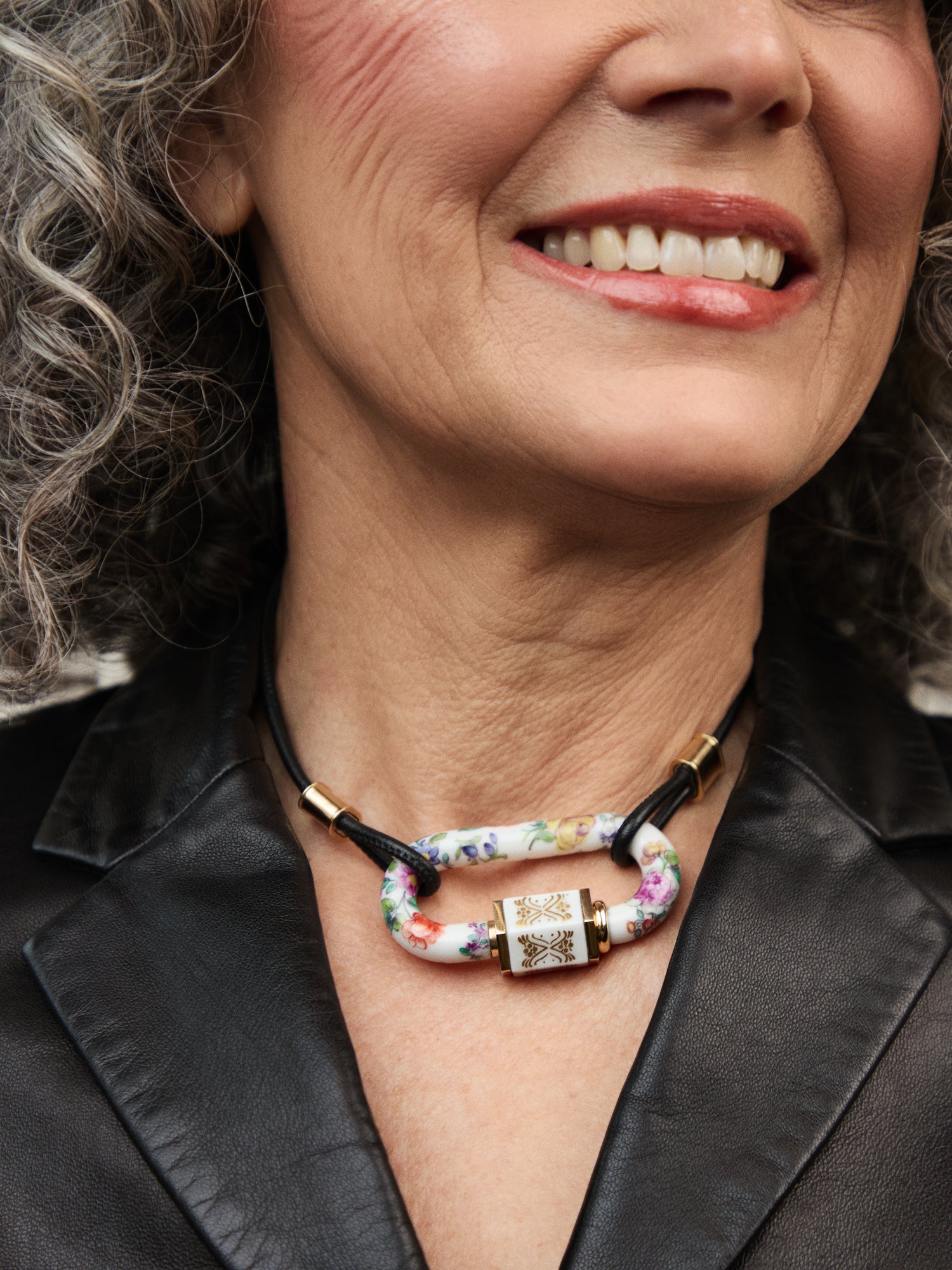Nahaufnahme einer Frau mit grauen Locken, die eine graue Bluse trägt und eine auffällige Halskette mit einem porzellanverzierten Verschluss in floraler Bemalung trägt.
Close-up of a woman with curly gray hair, wearing a gray blouse and a striking necklace with a porcelain clasp decorated with floral painting.