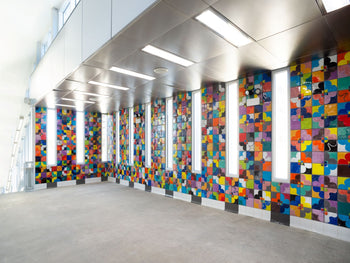 Farbenfrohe Wandinstallation mit glänzenden, handgefertigten Keramikfliesen in geometrischen Blumenmustern von Judy Ledgerwood im modernen, lichtdurchfluteten Gebäudeflur
Colorful wall installation featuring glossy, handcrafted ceramic tiles with geometric floral patterns by Judy Ledgerwood in a bright, modern corridor