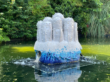 Große weiß und blau glasierte Keramikskulptur von Justin Lieberman als Brunnen in einem Wasserbecken, umgeben von dichtem Grün.

Large white and blue glazed ceramic fountain sculpture by Justin Lieberman standing in a reflecting pool, surrounded by lush greenery.