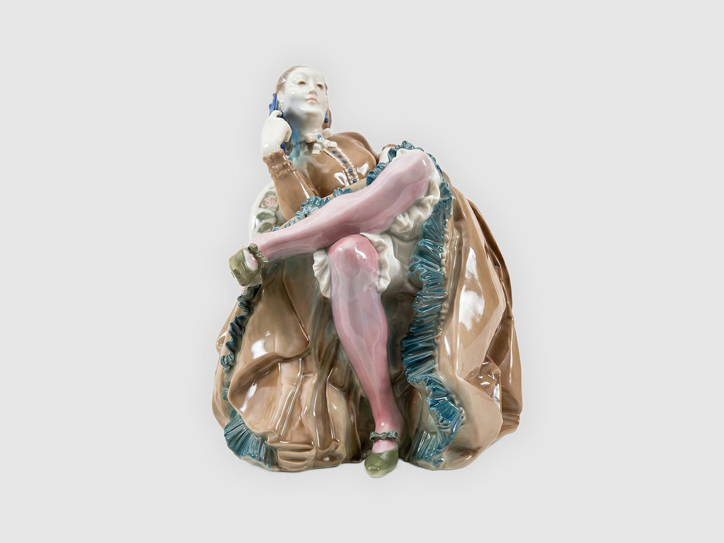 Frontale Ansicht der Porzellanfigur: Dame im Sessel mit übereinandergeschlagenen Beinen, rosa Strümpfen und reich gefälteltem braunem Kleid mit türkisfarbenen Rüschen.

Frontal view of the porcelain figurine: lady in an armchair with crossed legs, pink stockings and a richly pleated brown dress with turquoise ruffles.