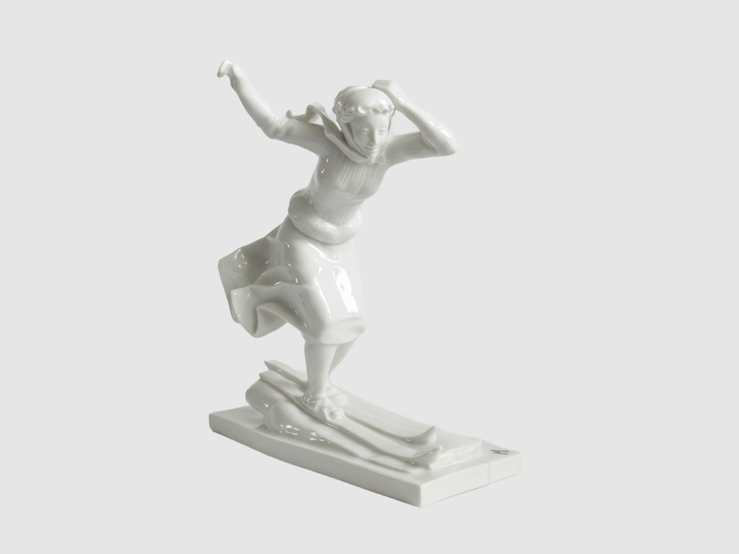 Weiße Porzellanfigur einer Skifahrerin in dynamischer Vorwärtsbewegung auf Skiern, mit flatterndem Rock und erhobenen Armen, auf rechteckigem Sockel.

White porcelain figurine of a female skier in dynamic forward motion on skis, skirt billowing and arms raised, on a rectangular base.