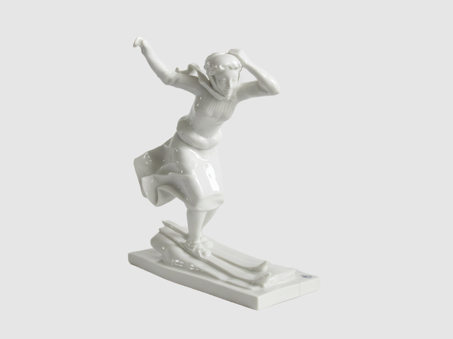 Weiße Porzellanfigur einer Skifahrerin in dynamischer Vorwärtsbewegung auf Skiern, mit flatterndem Rock und erhobenen Armen, auf rechteckigem Sockel.

White porcelain figurine of a female skier in dynamic forward motion on skis, skirt billowing and arms raised, on a rectangular base.