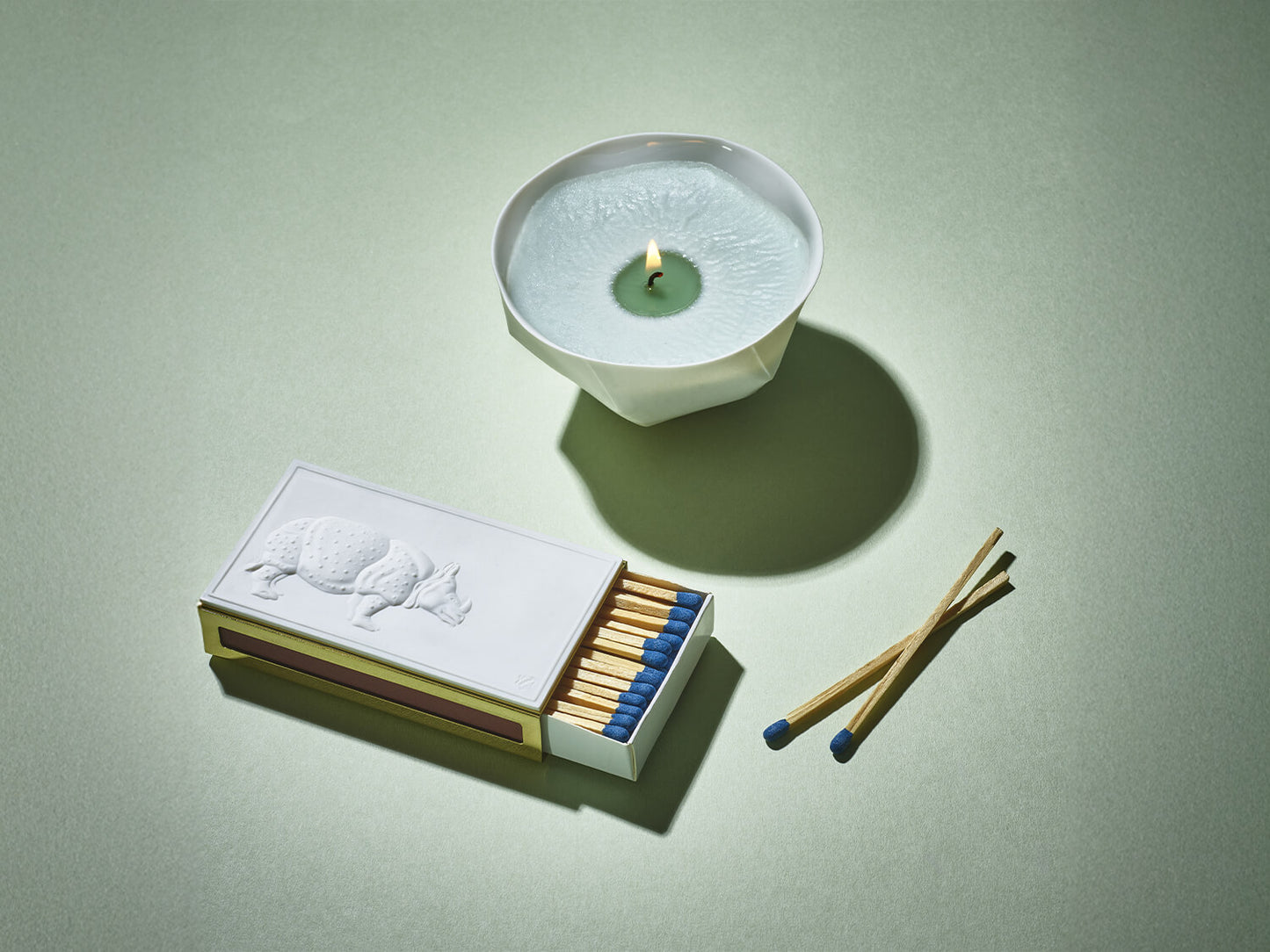 Streichholzetui mit Porzellanrelief und brennender Schale aus weißem Porzellan mit hellgrüner Kerze auf grünem Hintergrund.

Matchbox case with porcelain relief and a burning white porcelain bowl candle on a soft green background.