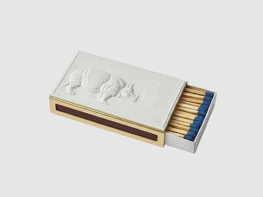 Streichholzetui mit weißer Porzellanplatte und Nashornrelief auf messingfarbener Hülle, Streichhölzer mit blauen Köpfen vor hellem Hintergrund.

Matchbox case with white porcelain plate and rhino relief on a brass-coloured sleeve, blue-tipped matches against a light background.