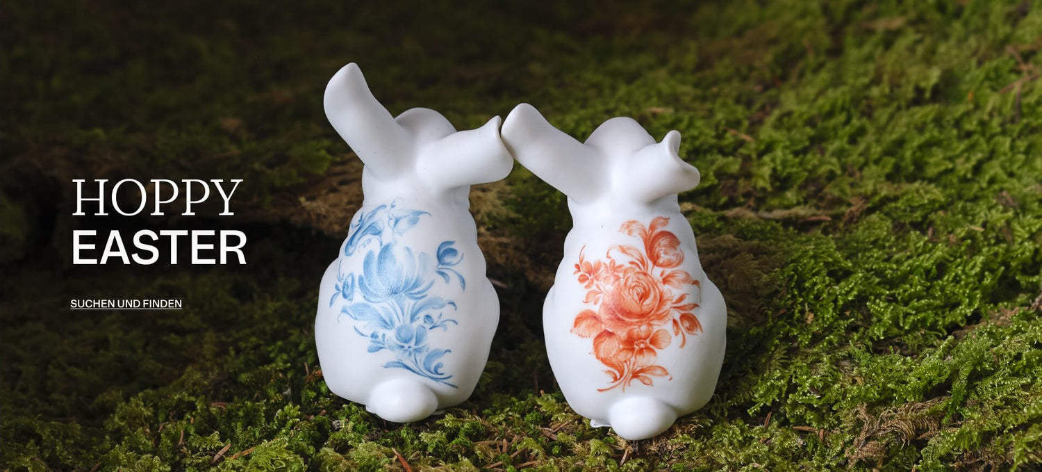 Zwei weiße Porzellanhasen mit blauer und korallroter Blumenmalerei stehen auf moosigem Waldboden; links ist der Schriftzug „HOPPY EASTER“ mit dem Linktext „SUCHEN UND FINDEN“ eingeblendet.

Two white porcelain rabbits with blue and coral floral decoration stand on a moss-covered forest floor; on the left, the words “HOPPY EASTER” and the link text “SUCHEN UND FINDEN” are overlaid