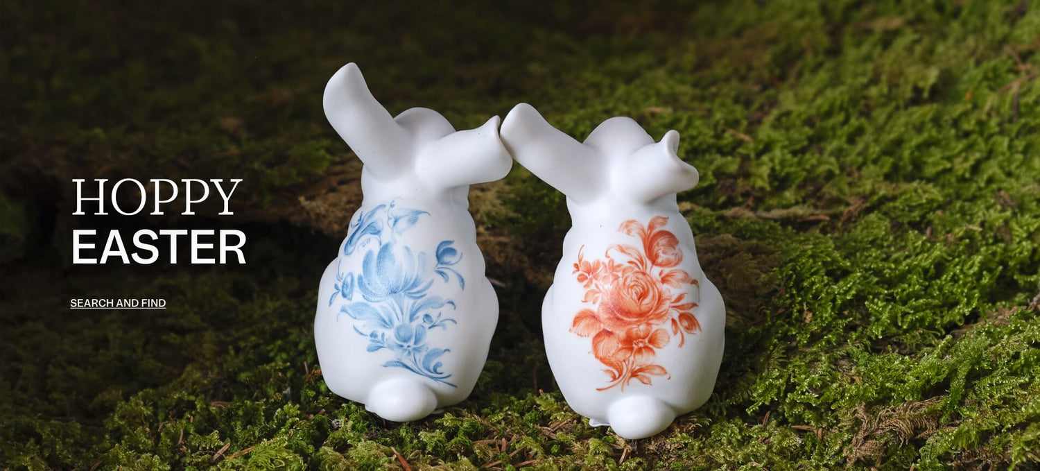 Zwei weiße Porzellanhasen mit blauer und korallroter Blumenmalerei stehen auf moosigem Waldboden; links ist der Schriftzug „HOPPY EASTER“ mit dem Linktext „SEARCH AND FIND“ eingeblendet.

Two white porcelain rabbits with blue and coral floral decoration stand on a moss-covered forest floor; on the left, the words “HOPPY EASTER” and the link text “SEARCH AND FIND” are overlaid.
